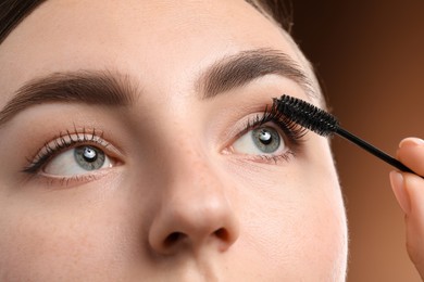 Photo of Young woman applying mascara onto her eyelashes on brown background, closeup
