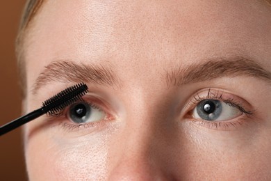 Photo of Young woman applying mascara onto her eyelashes on brown background, closeup