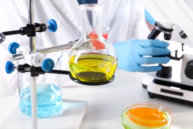 Photo of Scientist working with microscope at table indoors, focus on laboratory glassware with color liquids. Solution chemistry