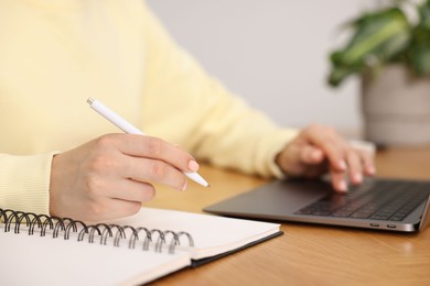 Photo of Copywriter using laptop and writing in notebook at wooden table indoors, closeup