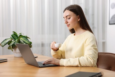 Photo of Copywriter with cup of drink using laptop at wooden table indoors