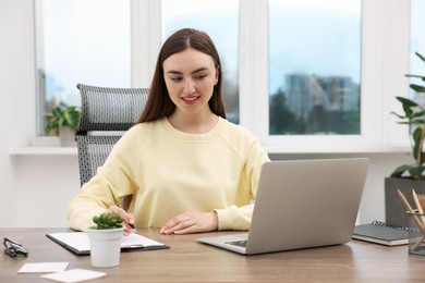 Photo of Copywriter using laptop and writing in notebook at wooden table indoors