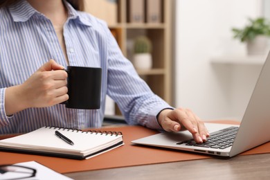 Photo of Copywriter with cup of drink using laptop at wooden table indoors, closeup