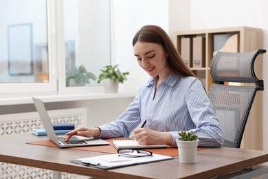 Photo of Copywriter using laptop and writing in notebook at wooden table indoors