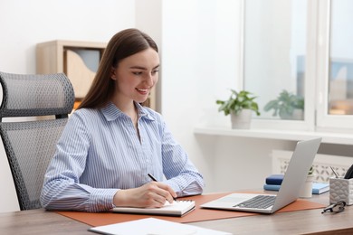 Photo of Copywriter using laptop and writing in notebook at wooden table indoors