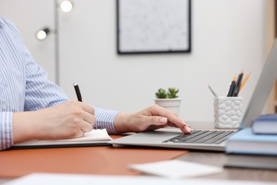 Photo of Copywriter using laptop and writing in notebook at wooden table indoors, closeup