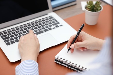 Photo of Copywriter with laptop writing in notebook at table indoors, closeup