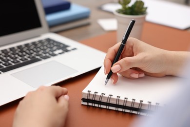 Photo of Copywriter with laptop writing in notebook at table indoors, closeup
