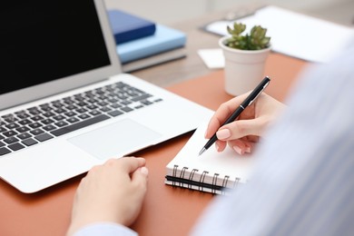 Photo of Copywriter with laptop writing in notebook at table indoors, closeup