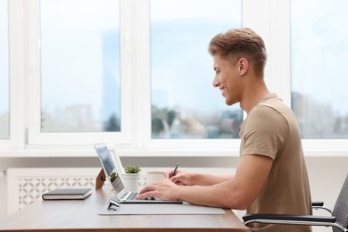 Photo of Copywriter using laptop and writing at wooden table indoors