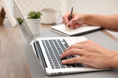 Photo of Copywriter using laptop and writing in notebook at table indoors, closeup