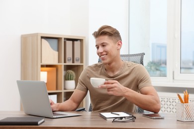 Photo of Copywriter with cup of drink using laptop at wooden table indoors