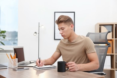 Photo of Copywriter with cup of drink and laptop writing in notebook at wooden table indoors