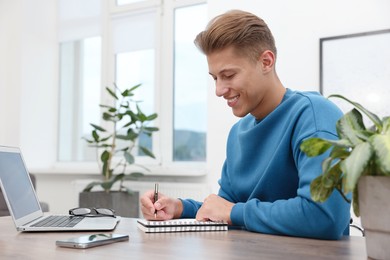 Photo of Copywriter with laptop writing in notebook at wooden table indoors