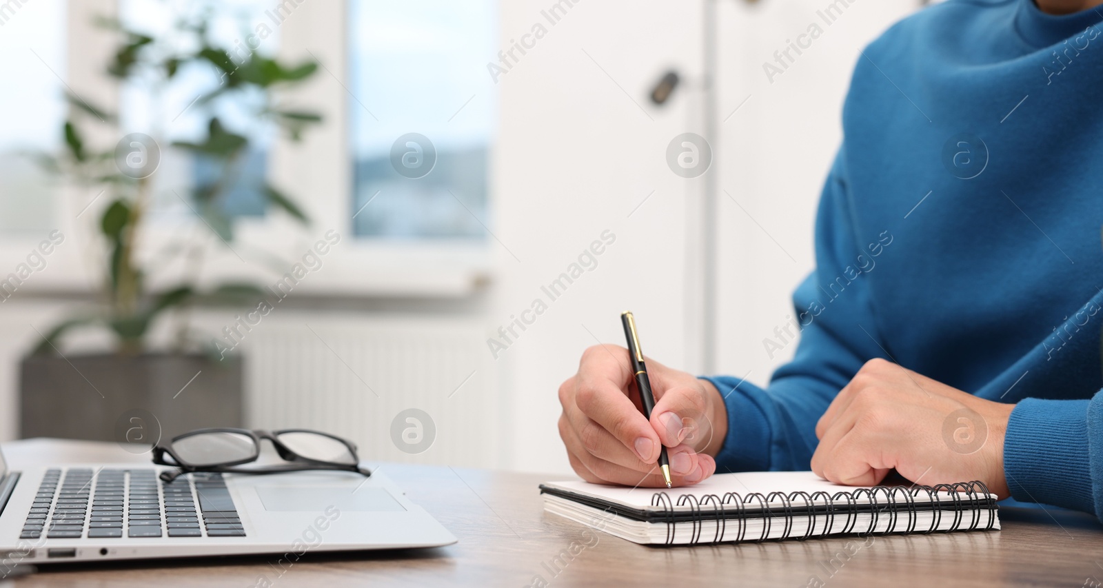 Copywriter with laptop writing in notebook at wooden table indoors, closeup Photo of Copywriter with laptop writing in notebook at wooden table indoors, closeup