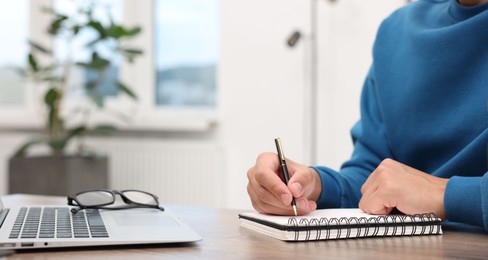 Photo of Copywriter with laptop writing in notebook at wooden table indoors, closeup
