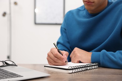 Photo of Copywriter with laptop writing in notebook at wooden table indoors, closeup