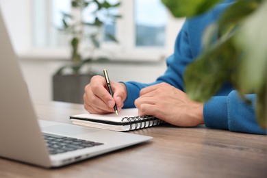 Photo of Copywriter with laptop writing in notebook at wooden table indoors, closeup
