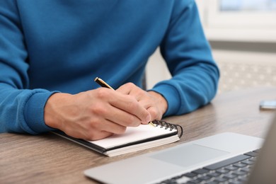 Photo of Copywriter with laptop writing in notebook at wooden table indoors, closeup