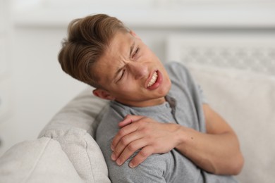 Photo of Young man suffering from shoulder pain on sofa at home