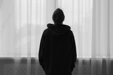 Photo of Silhouette of sad man near window at home, back view. Black and white effect
