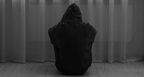 Photo of Sadness and depression. Silhouette of man in hoodie near curtains at home, back view. Black and white effect