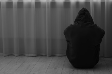 Photo of Sadness and depression. Silhouette of man in hoodie near curtains at home, back view and space for text. Black and white effect