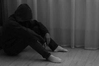 Photo of Sadness and depression. Man in hoodie near curtains at home. Black and white effect