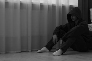 Photo of Sadness and depression. Man in hoodie near curtains at home, space for text. Black and white effect