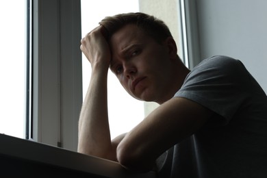 Photo of Sad thoughtful man near window indoors, low angle view. Depression