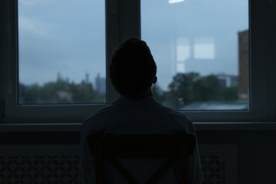 Photo of Sadness and depression. Silhouette of man near window at home, back view