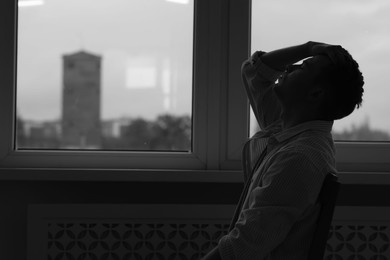 Photo of Sad tired man sitting on chair near window at home, space for text. Black and white effect