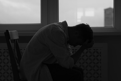 Photo of Sad man sitting on chair near window at home. Black and white effect