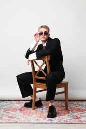 Photo of Handsome young man wearing stylish suit and sunglasses sitting on chair against white background