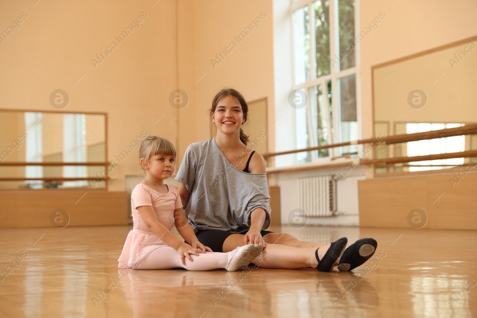 Photo of Little girl and her teacher sitting on floor in barre studio