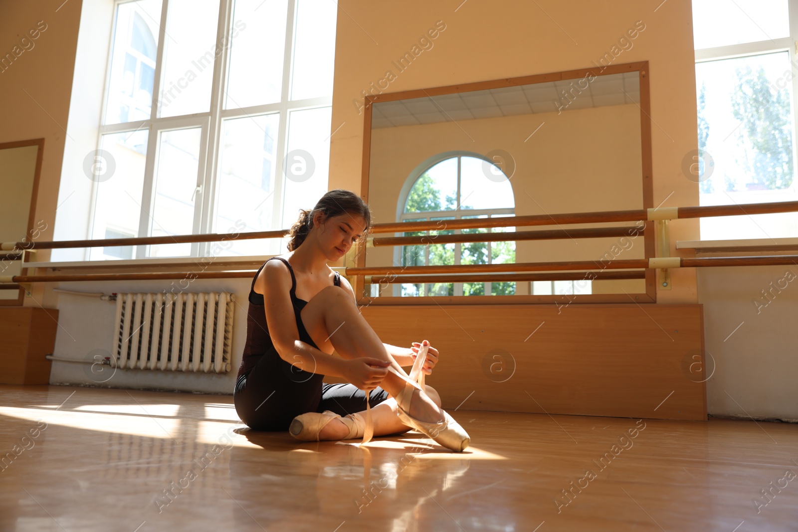 Photo of Beautiful teenage girl tying pointe shoes on floor in light barre studio