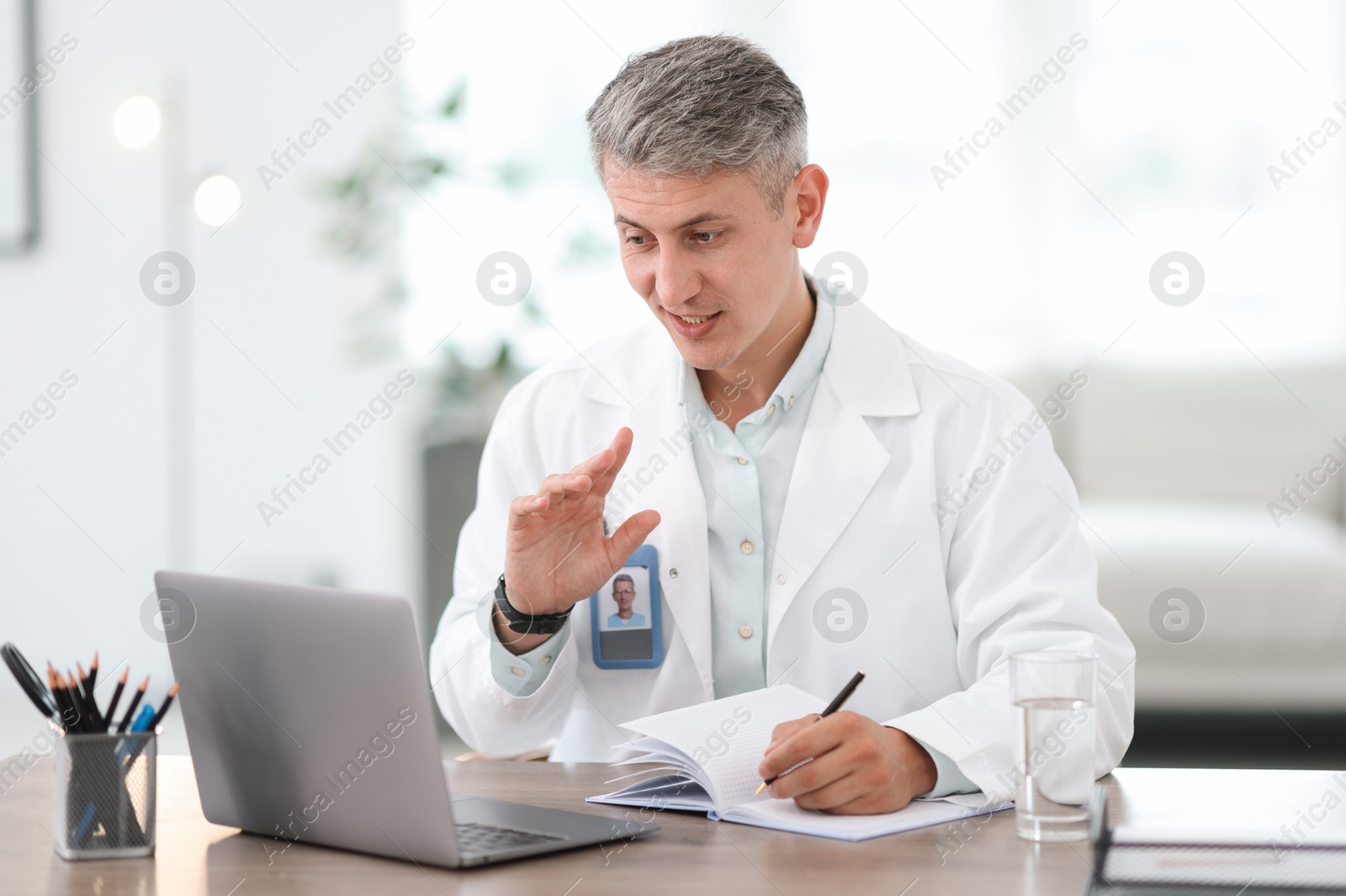 Photo of Telemedicine. Doctor making notes while having video call with patient via laptop indoors