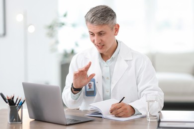 Photo of Telemedicine. Doctor making notes while having video call with patient via laptop indoors