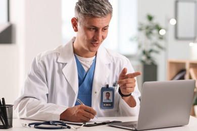 Photo of Telemedicine. Doctor making notes while having video call with patient via laptop indoors