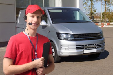 Image of Car service worker with headset and clipboard against vehicle outdoors. Client support