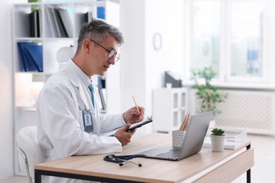 Photo of Telemedicine. Doctor making notes while having video call with patient via laptop indoors