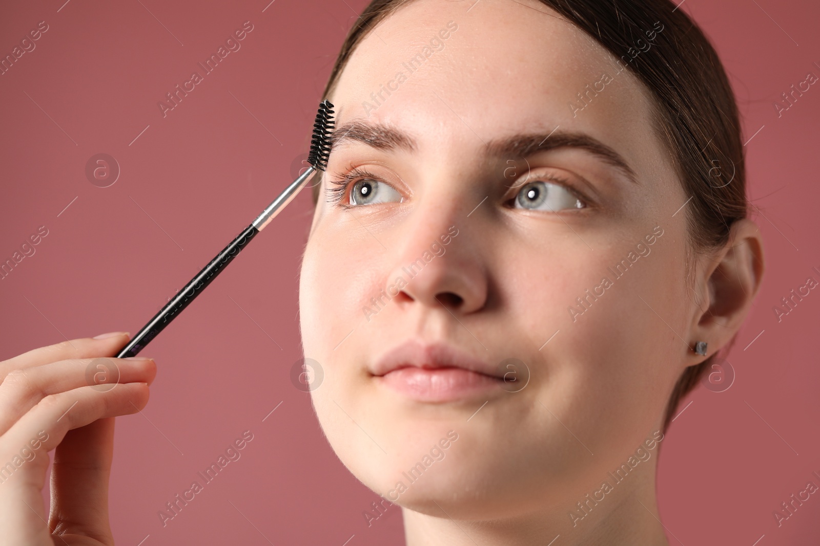 Photo of Young woman brushing her eyebrow with spoolie on pink background, closeup