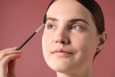 Photo of Young woman brushing her eyebrow with spoolie on pink background, closeup