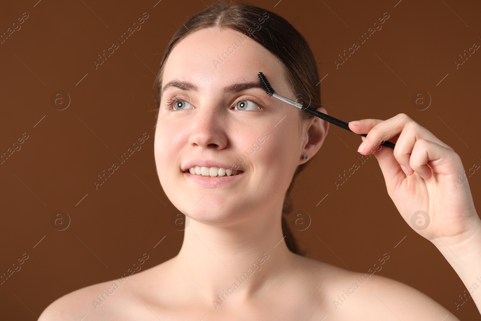 Photo of Young woman brushing her eyebrow with spoolie on brown background