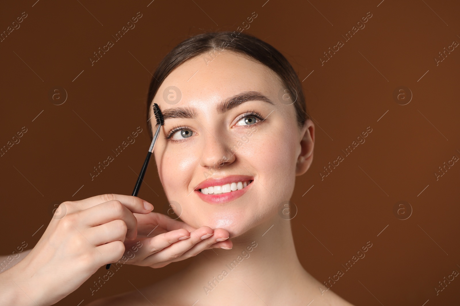 Cosmetologist brushing young woman's eyebrow with spoolie on brown background, closeup Photo of Cosmetologist brushing young woman's eyebrow with spoolie on brown background, closeup