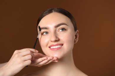 Photo of Cosmetologist brushing young woman's eyebrow with spoolie on brown background, closeup
