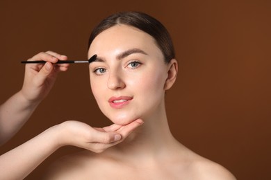 Photo of Cosmetologist brushing young woman's eyebrow with spoolie on brown background, closeup