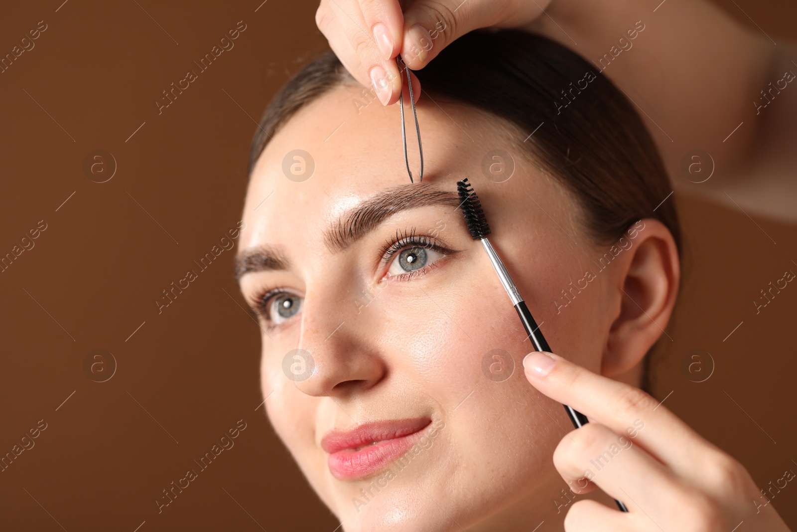 Photo of Cosmetologist plucking and brushing young woman's eyebrow on brown background, closeup