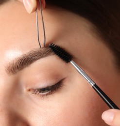 Photo of Cosmetologist plucking and brushing young woman's eyebrow, closeup