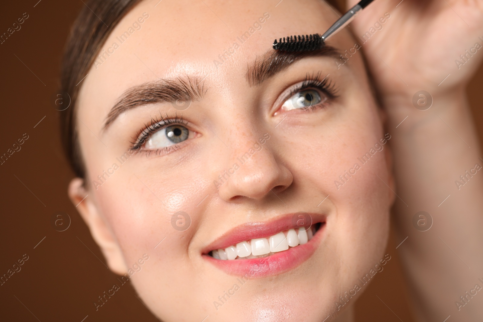 Cosmetologist brushing young woman's eyebrow with spoolie on brown background, closeup Photo of Cosmetologist brushing young woman's eyebrow with spoolie on brown background, closeup